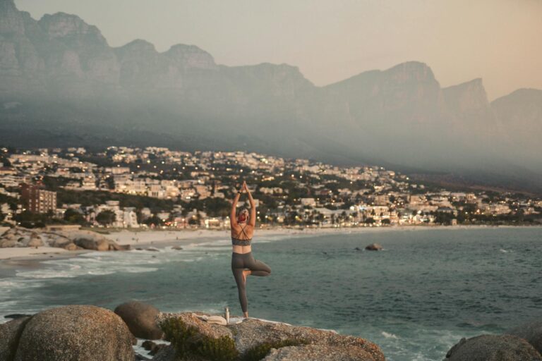 A person practices yoga on the rocks overlooking the sea and city lights during sunset in Cape Town.